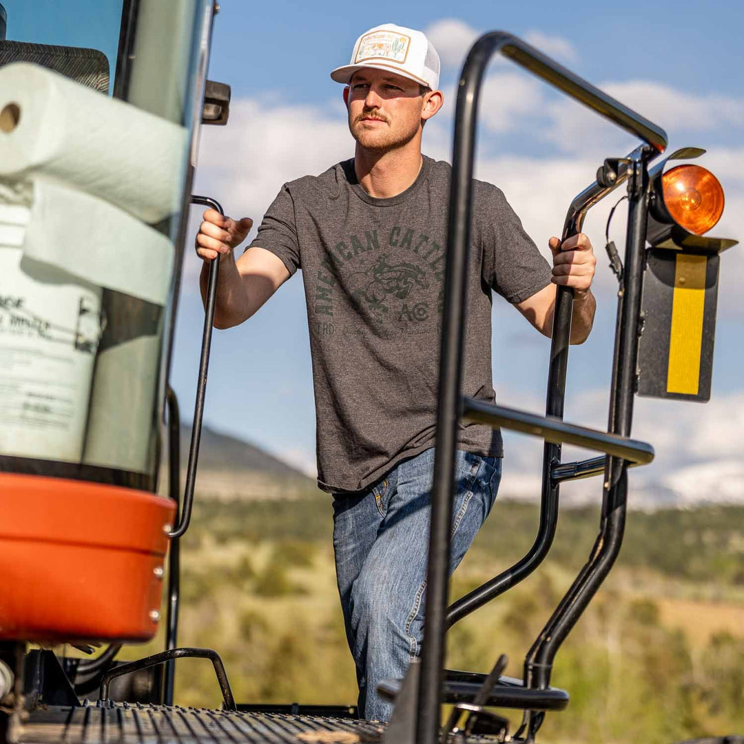 Wearing the bold Ride 'Em Cowboy Tee by American Cattle Co, jeans, and a white cap, a man stands on a large vehicle's step, gripping both handrails as rugged mountains behind him evoke an adventurous cowboy spirit.