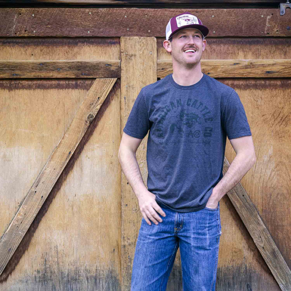 A man wearing a dark American Cattle Co Ride 'Em Cowboy Tee, blue jeans, and a trucker hat stands before a large wooden barn door, gazing to the side with one hand in his pocket and the other at his side.