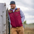 A man in Rural Cloth's Rider red quilted insulated vest, black cap, grey hoodie, and brown pants leans against a wooden post outdoors, smiling with a cloudy sky and blurred landscape behind him.
