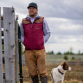 A man in the Rural Cloth Rider maroon quilted insulated vest, gray hoodie, brown pants, and black cap stands by a wooden fence outdoors with a brown and white dog against a cloudy sky and open field.