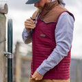 A person wearing the Rural Cloth Rider vest in maroon/brown over a gray hoodie zips it up while standing outdoors by a wooden post and metal gate. Their face is partially obscured by a cap.