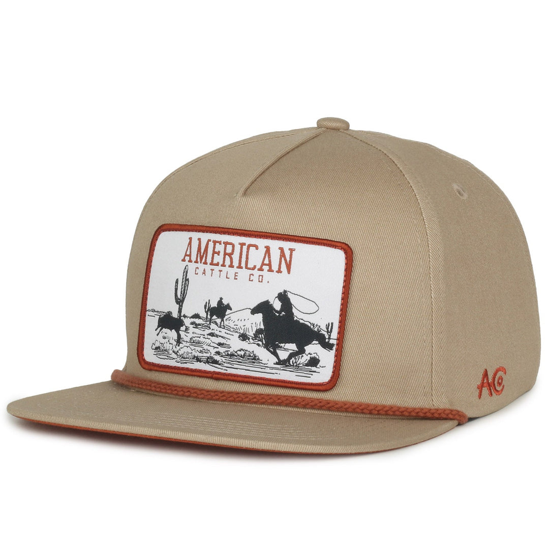 The Roper Hat by American Cattle Co is a beige baseball cap with Western flair, featuring a cowboy roping cattle among desert cacti and "American Cattle Co." at the top. A brown string along the brim completes this must-have for ranching accessory enthusiasts.
