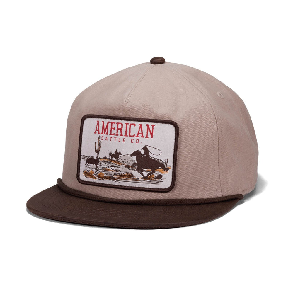 The Roper Hat by American Cattle Co features a tan and brown design with a front patch of a cowboy lassoing a calf, desert scenery, and "AMERICAN CATTLE CO." embroidered in red and brown.
