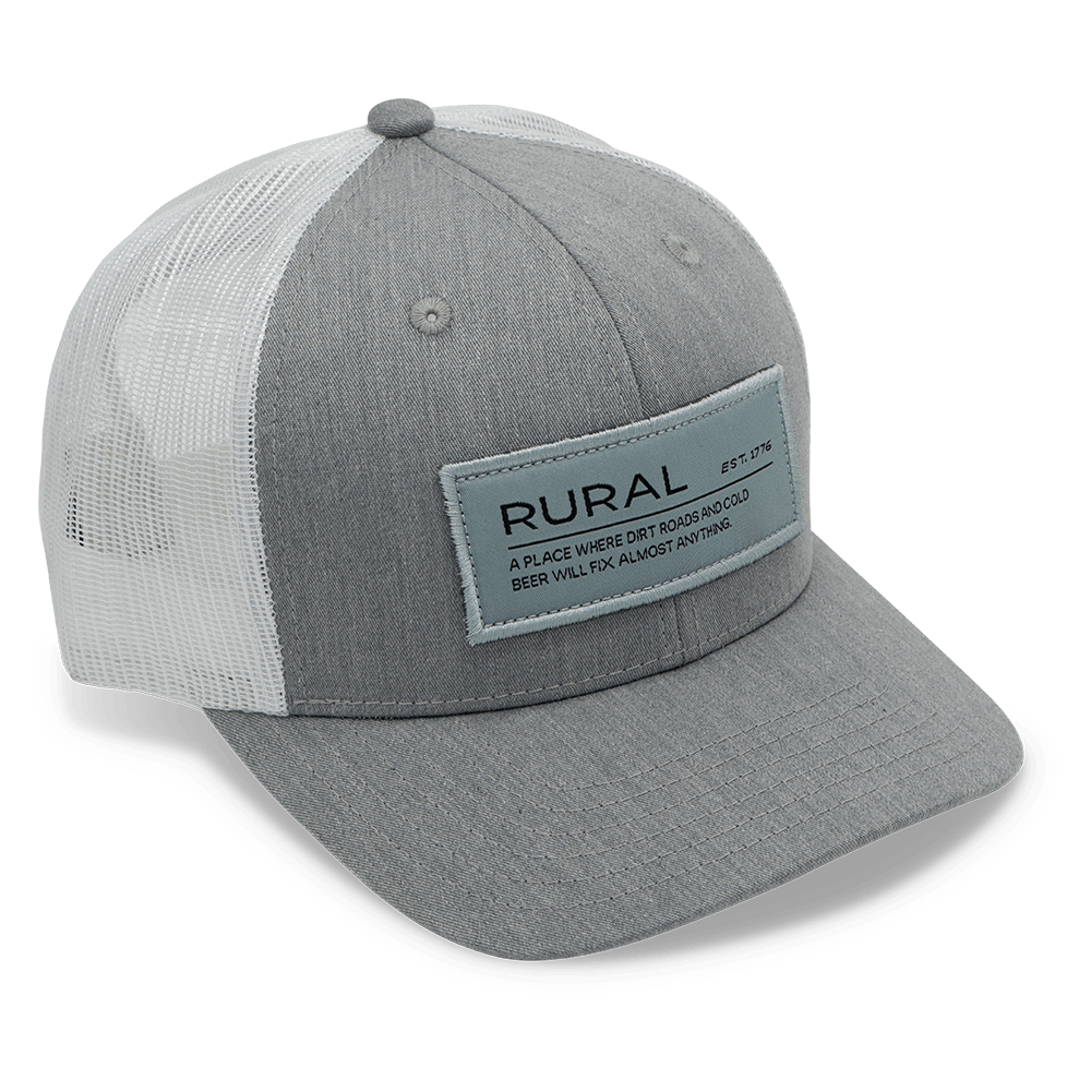 The American Cattle Co Rural Def Hat is a gray and white cap with a mesh back, curved brim, ventilation holes, adjustable snapback, and front patch reading "RURAL" above smaller text.