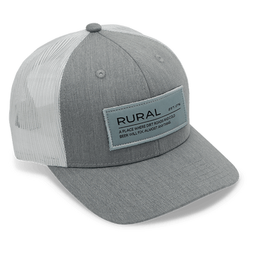 The American Cattle Co Rural Def Hat is a gray and white cap with a mesh back, curved brim, ventilation holes, adjustable snapback, and front patch reading "RURAL" above smaller text.