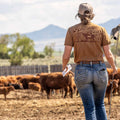 Wearing jeans, a cap, and the brown Shadowhorn Tee from Rural Cloth featuring a longhorn silhouette, a person stands holding papers and a stick while observing brown cattle in a mountain-fenced outdoor area.