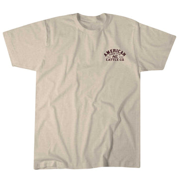 Introducing the Shadowhorn Tee by American Cattle Co: a beige T-shirt with "American Cattle Co." in dark red on the left chest, featuring a subtle longhorn silhouette. This flat-laid, short-sleeve shirt with a round neckline embodies classic Western style.