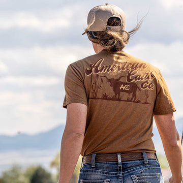 A person in a tan cap and a brown American Cattle Co Shadowhorn Tee, featuring a desert scene and longhorn silhouette on the back, stands outdoors facing away from the camera with a slightly blurred background.