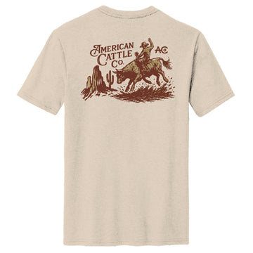 The Stampede Tee by American Cattle Co is beige, made from a soft cotton blend, and features a brown cowboy-on-bull graphic with cacti, desert scene, and the text "American Cattle Co. AC." on the back in a classic fit.