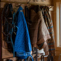 Three Rural Cloth Sunday Jackets—blue quilted, brown puffer, and plaid—hang on wooden hooks in a rustic setting with leather straps and horse tack also on the nearby walls.