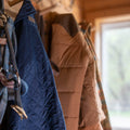 Several coats, including the Rural Cloth Sunday Jacket—a blue quilted shirt jacket with polar fleece lining—hang alongside a tan puffer on wooden hooks in a softly lit hallway.