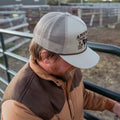 A man with a brown beard, wearing the Rural Cloth Cold Snap Puffer and a light mesh baseball cap, leans on a metal fence in an outdoor ranch setting. His face is mostly turned downward and obscured.