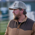 A bearded man in Rural Cloth's beige and brown Cold Snap Puffer and a light trucker hat looks left outdoors, with a fence and blurred background behind him.