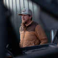 A bearded man in a baseball cap stands outdoors by a vehicle, wearing the tan and brown Cold Snap Puffer by Rural Cloth. A metal building can be seen in the background.