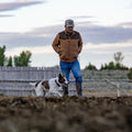A man wearing Rural Cloth's The Cold Snap Puffer, blue jeans, and boots walks outdoors with a brown and white dog. A wooden fence and trees stand in the background beneath a cloudy sky.