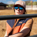 A person wears The Switchback Reversible Puffer by Rural Cloth, a tan and orange hat, blue reflective sunglasses, and a brown shirt while standing outdoors near a metal fence on a sunny day.