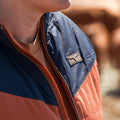 A person wears The Switchback Reversible Puffer by Rural Cloth, featuring orange and navy panels with a western yoke and chest logo, while standing outdoors against a blurred brown background.