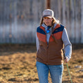 A person wearing The Switchback Reversible Puffer by Rural Cloth stands outdoors holding a stick, with a blurred wooden fence in the background.