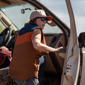 A person in Rural Cloth's The Switchback Reversible Puffer stands by an open car door on a sunny day, wearing a hat and sunglasses, looking away from the vehicle.