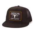 The Trademark Hat by American Cattle Co is a black snapback trucker cap with a mesh back and flat brim. The front showcases a rectangular patch with a white longhorn graphic in the center, framed by a gold border, and 