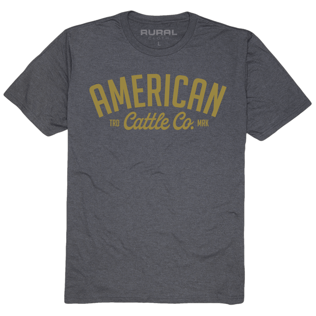 The Rural Cloth Trademark Tee is a heather gray shirt featuring "AMERICAN Cattle Co." in bold gold letters and "TRD" and "MRK" flanking the design, crafted from premium weight fabric for comfort and durability.