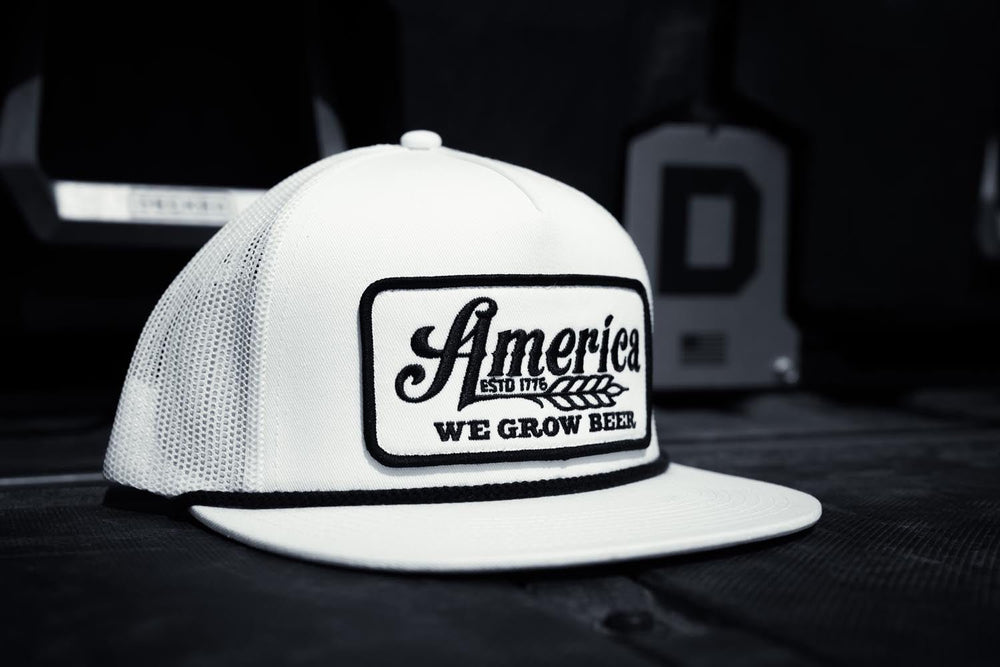 The American Cattle Co We Grow Beer Flat is a white mesh back snapback hat with a flat bill and a patch that reads “America ESTD 1776 WE GROW BEER” in black lettering, shown resting on a dark surface with blurred objects behind it.
