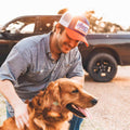A man wearing a gray button-up shirt and a We Grow Beer Hat in red, white, and blue is outdoors, petting a golden retriever. A black pickup truck is parked in the background with sunlight filtering through the trees, capturing an idyllic American scene reminiscent of summer days shared with a cold beer.