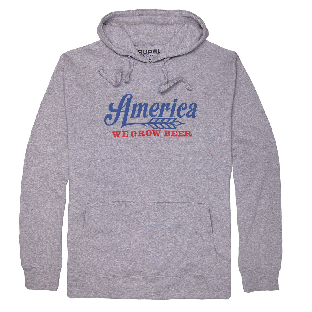 The We Grow Beer Pullover by American Cattle Co is a heather gray hoodie with "America" in blue above a wheat graphic and "WE GROW BEER" in bold red. Made from premium anti-pill fabric, it features an ultra-soft interior, front pocket, and drawstrings.