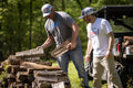 Two men, dressed in hats and casual clothing, are stacking firewood outdoors. One man is placing a piece of wood on the pile while the other watches, holding a Coors Light. Nearby, a vehicle is parked in a grassy, wooded area with cups and a can of Silver Bullet resting on its hood. One of the men is wearing a We Grow Coors Light Tee from Rural Cloth.