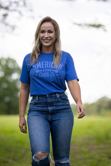 A woman smiles outdoors on grass, wearing the American Cattle Co Western Tee with high-waisted ripped jeans. Trees and greenery blur in the background, highlighting her casual nod to classic Western style.