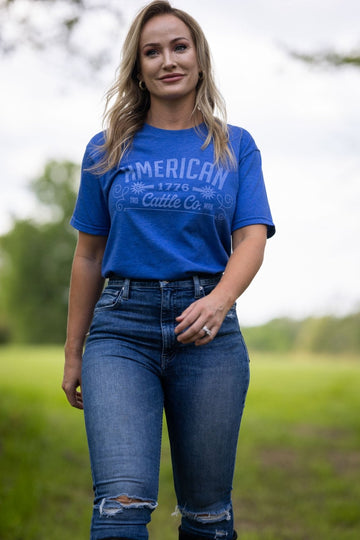 A woman with long blonde hair walks outdoors on grass, wearing the American Cattle Co Western Tee and distressed blue jeans. "American 1776 Cattle Co." is printed on the tee, celebrating rural style with trees and a cloudy sky in the background.