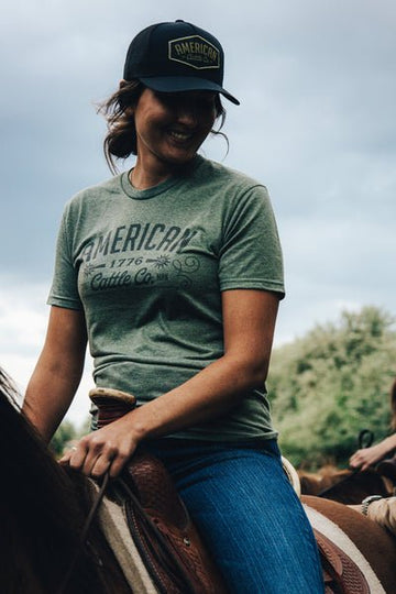 Someone wearing an American Cattle Co Western Tee, blue jeans, and a black cap sits on a horse outdoors beneath a cloudy sky, reflecting the rugged American cattle industry spirit.