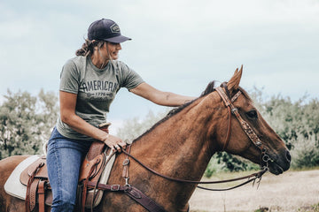 Wearing an American Cattle Co Western Tee, cap, and jeans, a rider sits on a brown horse outdoors, holding the reins and patting the horse’s neck. Trees and a cloudy sky set the scene for this tribute to American cattle culture.