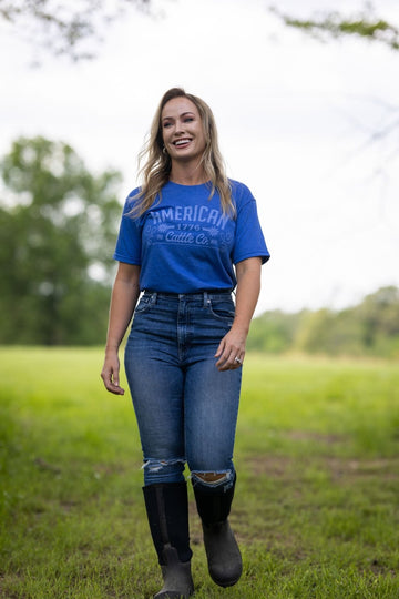 A woman with long blonde hair, wearing a blue Western Tee from American Cattle Co, jeans, and black boots walks on grass outdoors, with trees and a cloudy sky in the background.