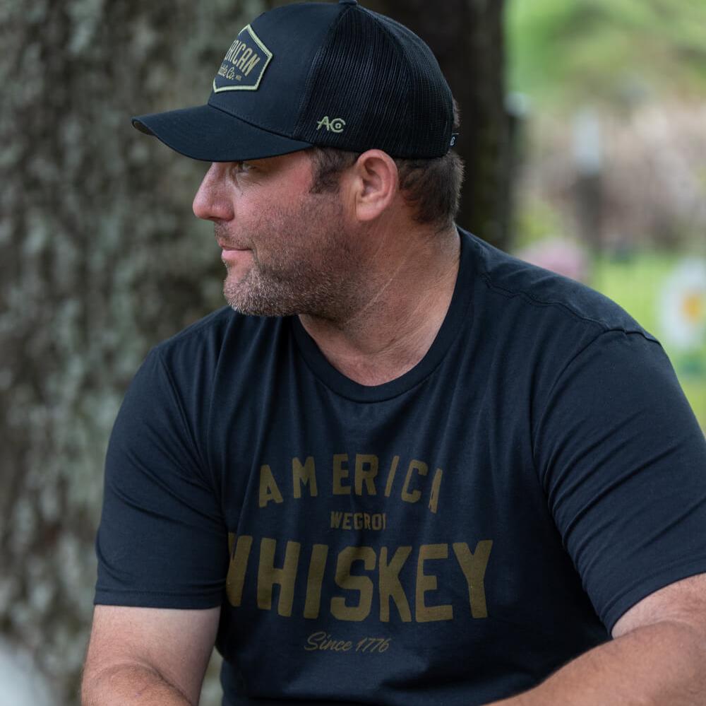 A whiskey lover sits outdoors in a Rural Cloth Whiskey Tee and black cap, turned sideways with a tree trunk behind him.