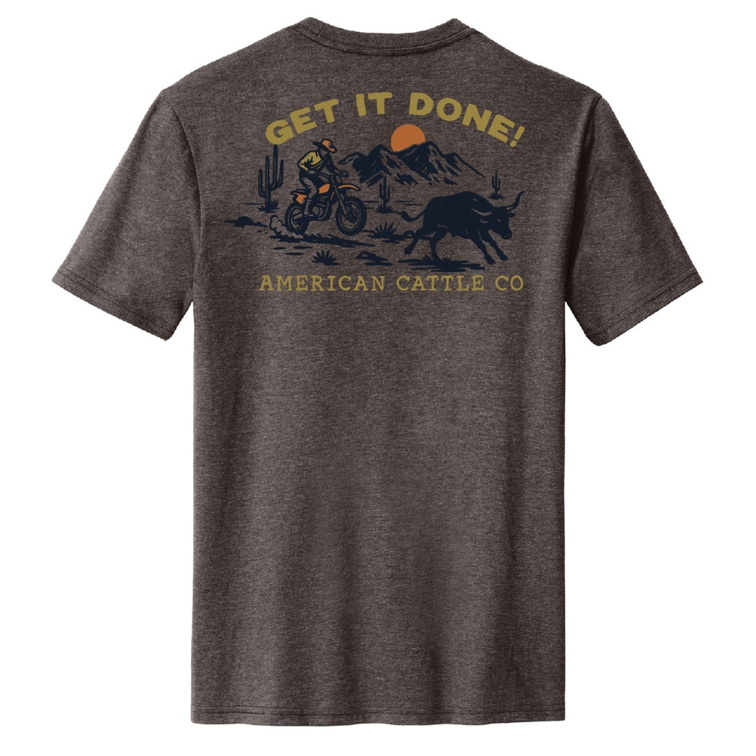 The Workhorse Tee by American Cattle Co features a dark gray vintage design of a dirt biker riding near a bull with mountains, cacti, and the sun. “GET IT DONE!” decorates the top; “AMERICAN CATTLE CO.” is printed below for rugged style.