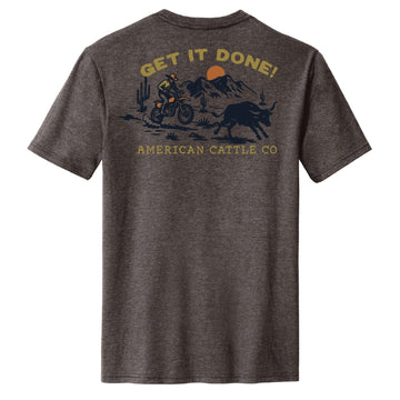 The Workhorse Tee by American Cattle Co features a dark gray vintage design of a dirt biker riding near a bull with mountains, cacti, and the sun. “GET IT DONE!” decorates the top; “AMERICAN CATTLE CO.” is printed below for rugged style.