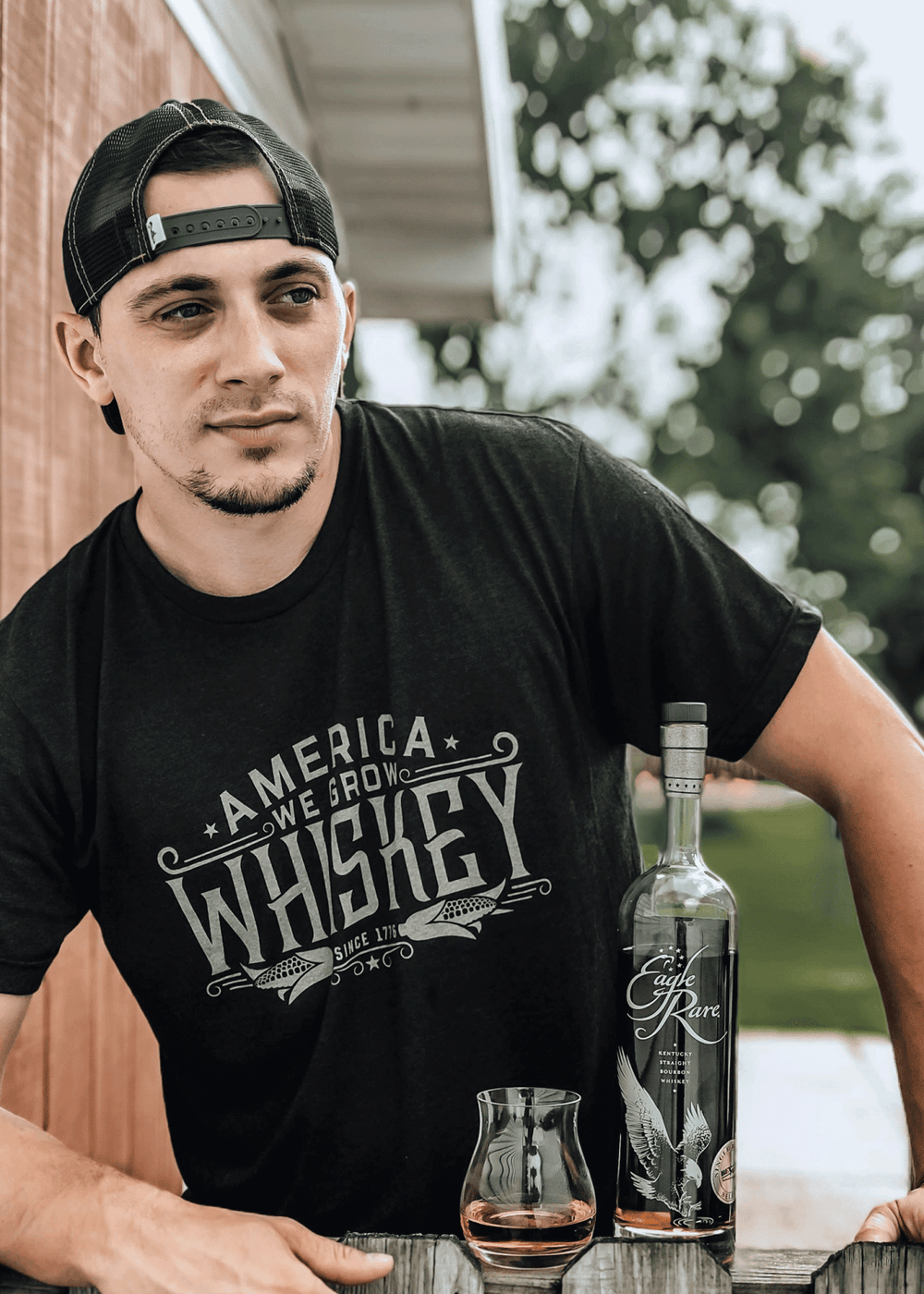 A man in a modern athletic fit America We Grow Whiskey Tee by Rural Cloth, cap worn backwards, stands beside a whiskey bottle and glass on a wooden outdoor surface.