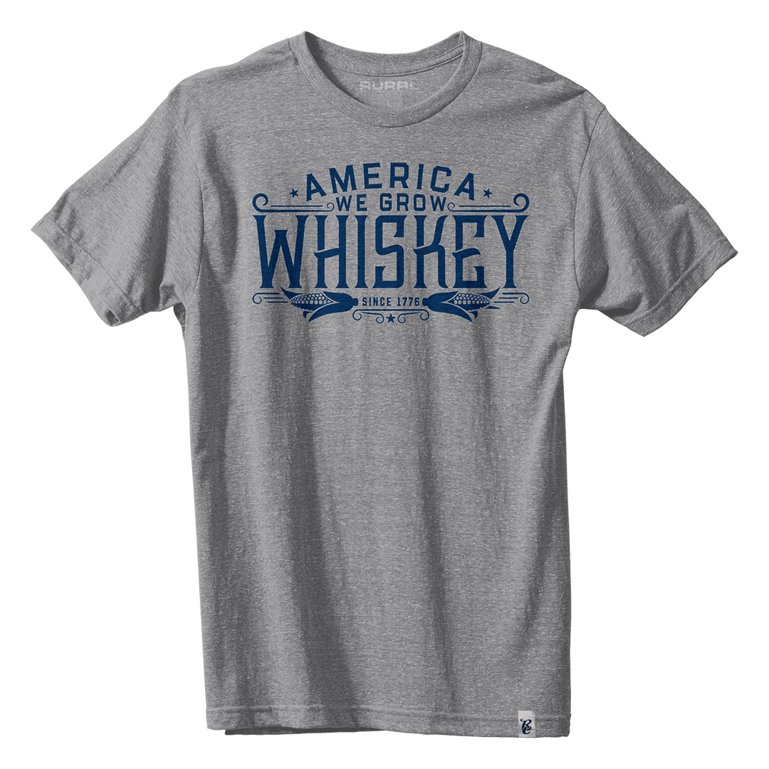 The America We Grow Whiskey Tee - Gray by American Cattle Co is a heather gray short-sleeve shirt with blue "America We Grow Whiskey Since 1776" text, decorative elements, and two wheat stalk illustrations. Made in the USA.