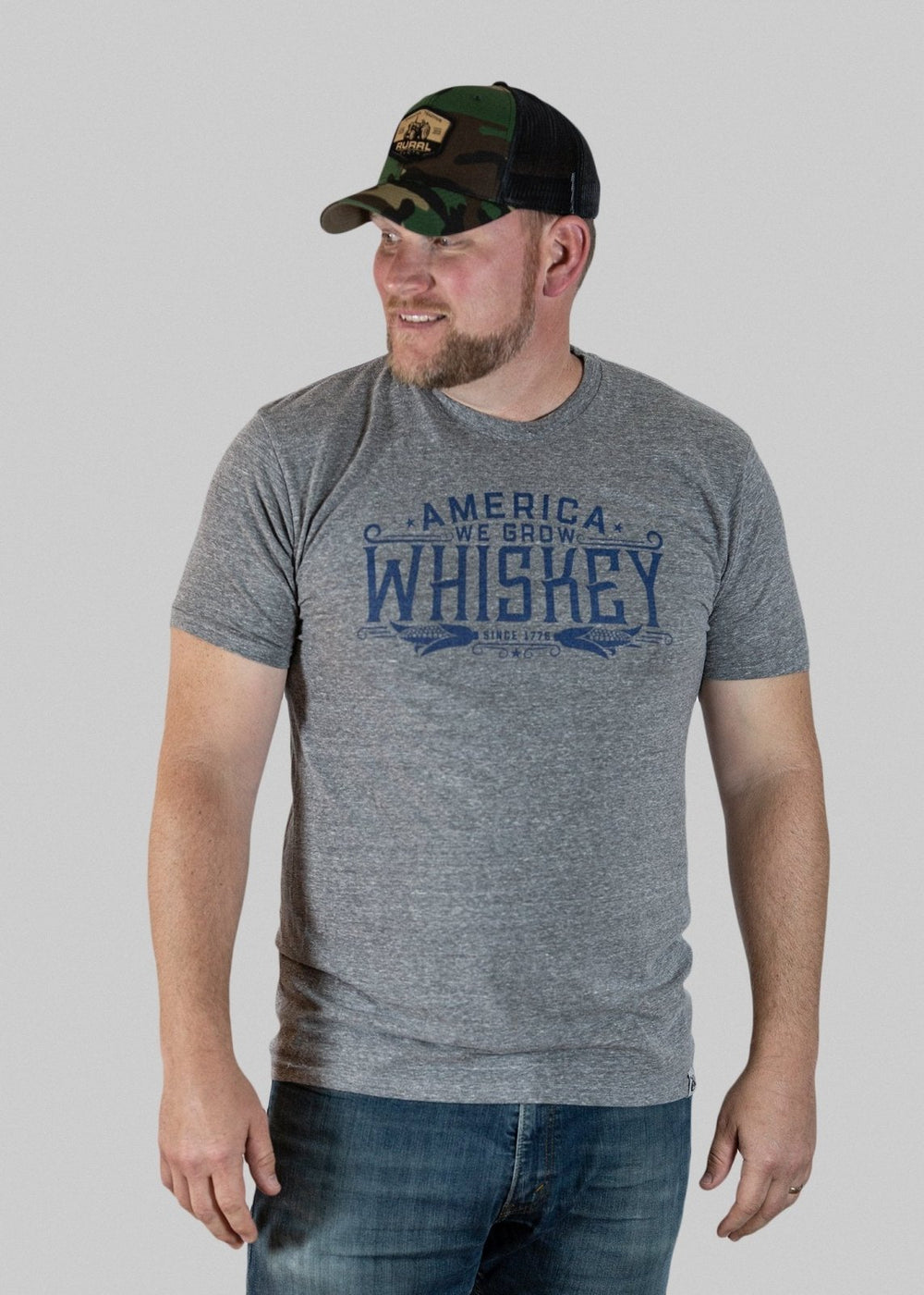 A man wears the American Cattle Co America We Grow Whiskey Tee - Gray and a camo cap, standing against a light gray background. He looks left and smiles, highlighting classic corn farmer style.