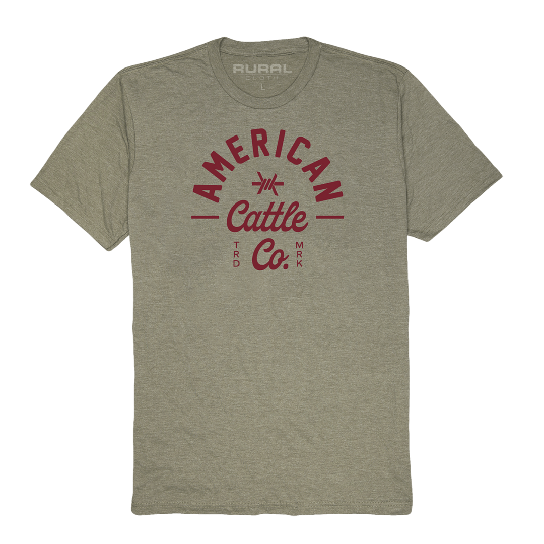 The Barb Tee by American Cattle Co is a heather gray short-sleeve t-shirt in a premium cotton blend, with bold red "American Cattle Co. TRD MRK" chest text and subtle "RURAL" detail inside the collar—a classic rancher style.