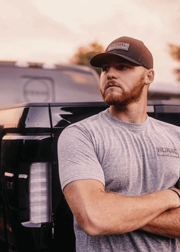A man with a reddish beard, wearing a gray t-shirt and a Rural Cloth Brandmark Hat, stands with arms crossed before a black truck, gazing to the side against a softly lit sky.