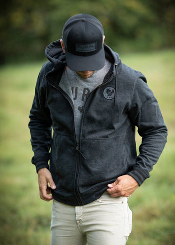 A person in a black hoodie with an embroidered rural logo, gray t-shirt, light pants, and a Brandmark Hat from Rural Cloth stands on grass outdoors, looking down while adjusting the hoodie. Trees blur in the background.