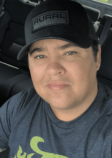 A person with short dark hair sits in a car, wearing a Rural Cloth Brandmark Hat and a dark T-shirt. They are looking at the camera with a neutral expression.