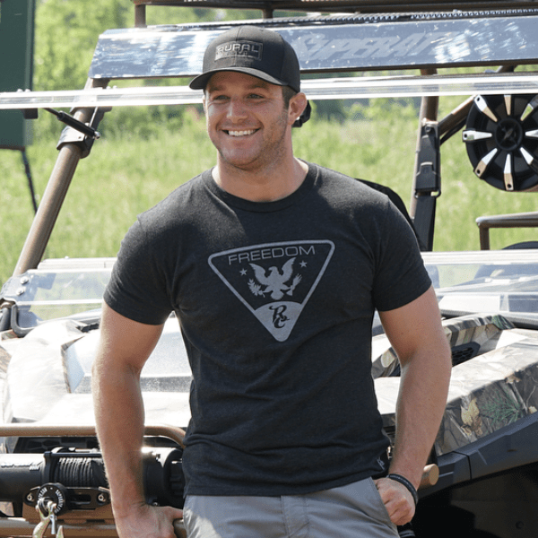 A man wearing a black Rural Cloth Brandmark Hat and a dark "Freedom" graphic tee stands smiling with one hand in his pocket before an off-road vehicle, with green grass and trees in the background.