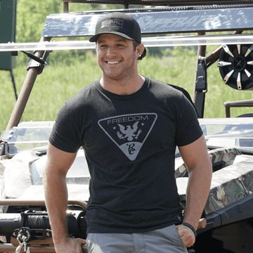A man wearing a black Rural Cloth Brandmark Hat and a dark "Freedom" graphic tee stands smiling with one hand in his pocket before an off-road vehicle, with green grass and trees in the background.
