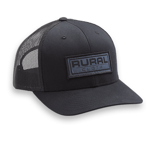 The Brandmark Hat by Rural Cloth is a black mesh trucker hat with a curved bill, structured front panel, adjustable snapback closure, and a rectangular patch embroidered with the Rural logo.