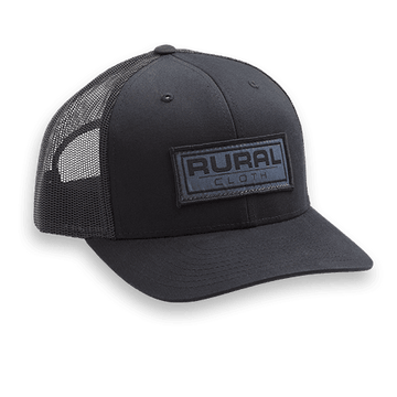 The Brandmark Hat by Rural Cloth is a black mesh trucker hat with a curved bill, structured front panel, adjustable snapback closure, and a rectangular patch embroidered with the Rural logo.