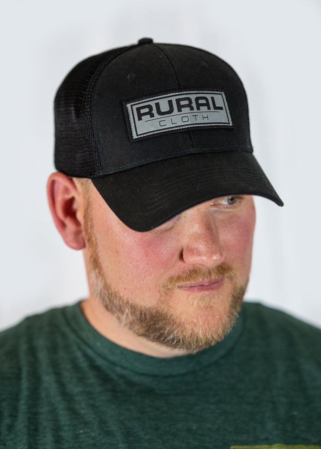 A man with a short beard wears a dark green shirt and the Rural Cloth Brandmark Hat, an adjustable black mesh baseball cap with a rectangular "RURAL CLOTH" patch. The background is plain white.