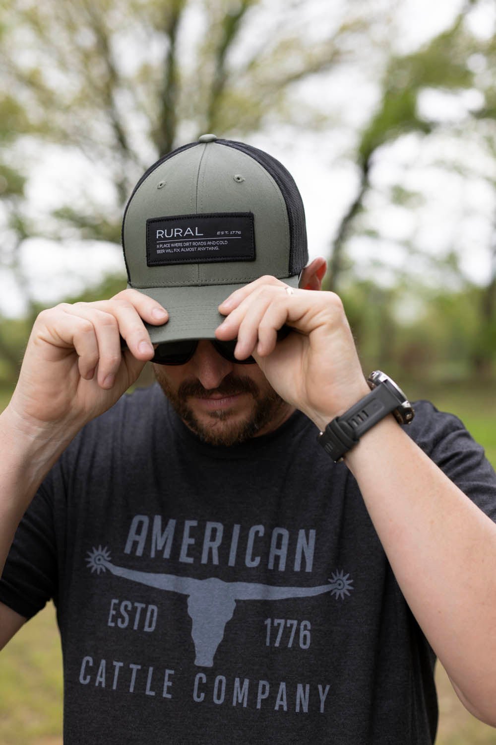 Wearing a black Bull Spurs Tee-Black Frost and a gray hat with a “Rural” patch, a person adjusts their black smartwatch outdoors, the trees softly blurred in the background.